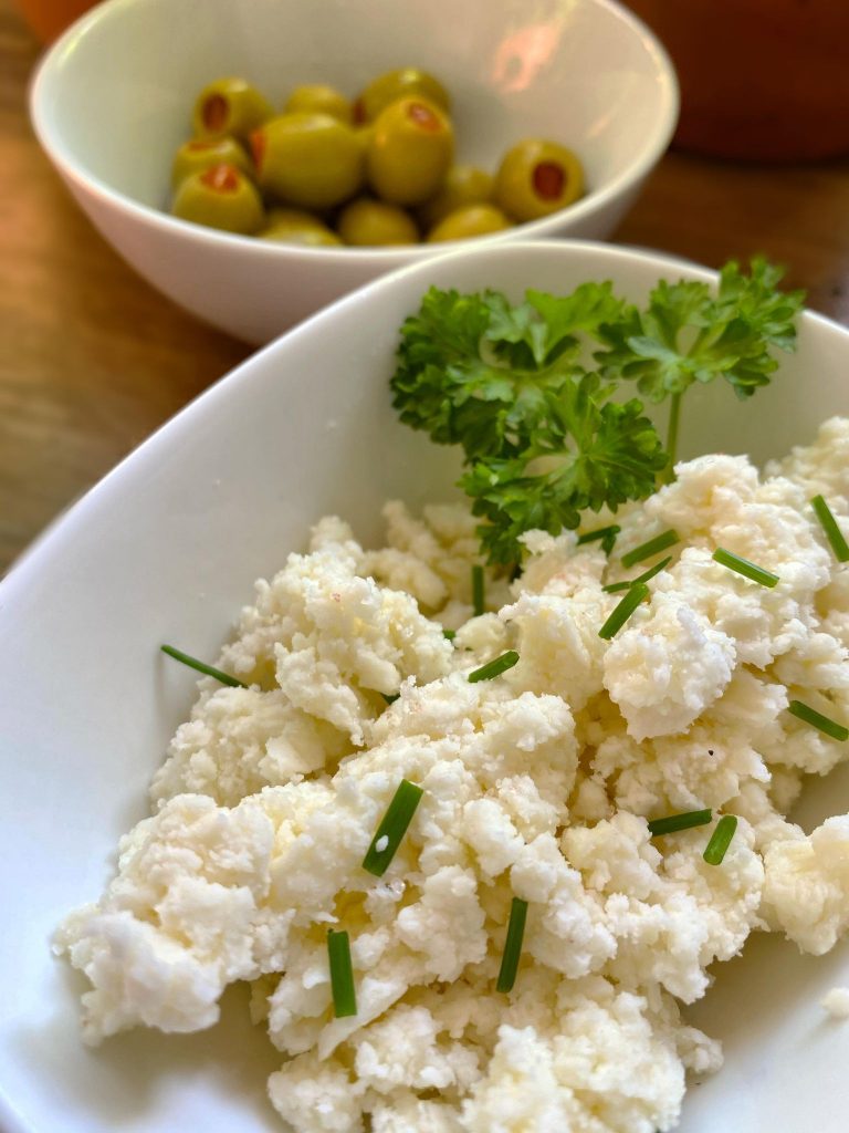 bowl with raw milk cottage cheese with chopped chives and sprig of parsley and bowl with olives in background