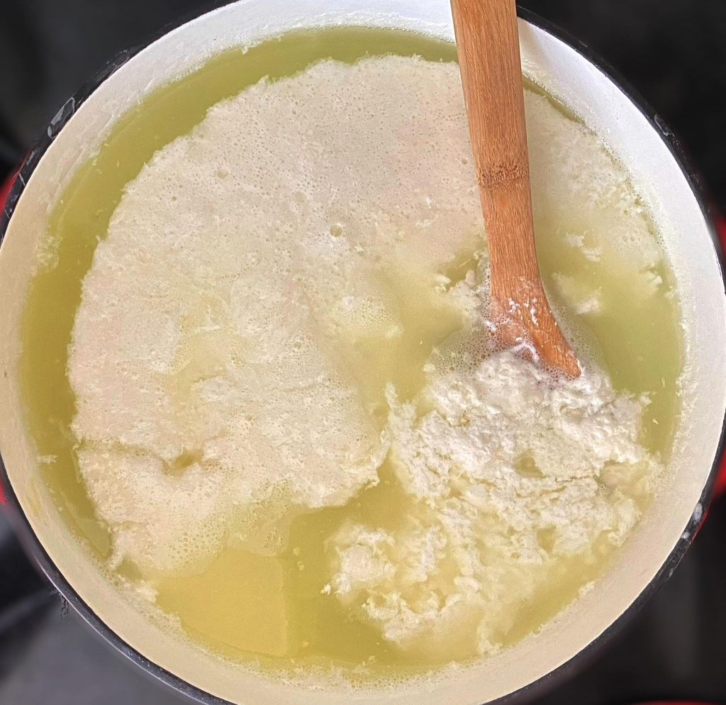 large pot filled with whey and separated milk curds with wooden spoon submerged in milk.
